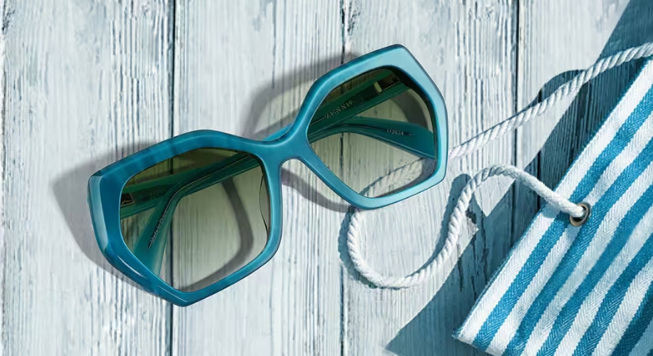 Blue hexagonal sunglasses with green lenses on a light wooden surface, next to a blue and white striped bag.