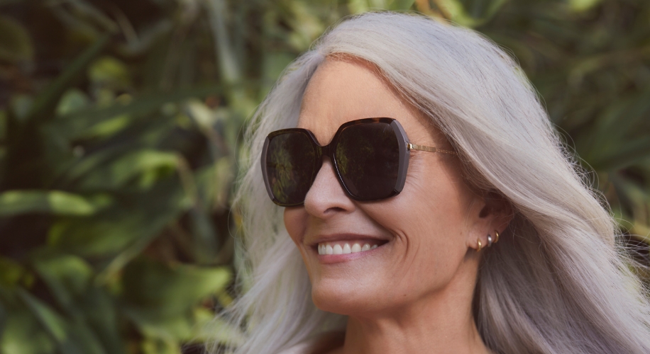 Woman smiling while wearing large, square-shaped, dark sunglasses with gold detailing on the arms.