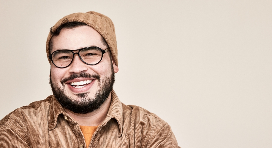 Man wearing brown corduroy jacket, mustard knit beanie, and black-framed glasses smiling.