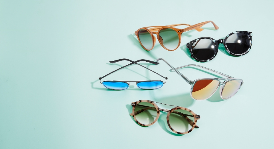 Various styles of sunglasses in black, brown, silver, and tortoiseshell frames on a light blue background.