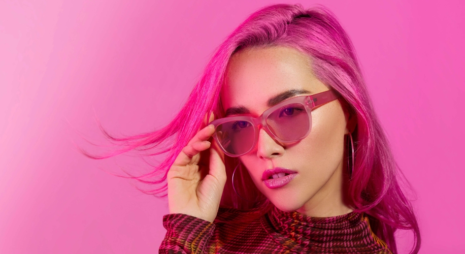 Woman with pink hair wearing pink-tinted sunglasses and a patterned top against a pink background.