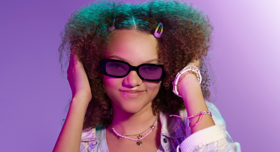 Girl wearing black sunglasses, colorful hair clips, and multiple bracelets, smiling against a purple gradient background.