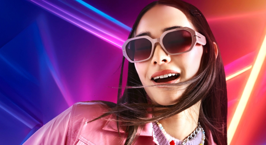 Woman wearing pink sunglasses with gradient lenses, dressed in a pink jacket, with glowing neon lights behind.