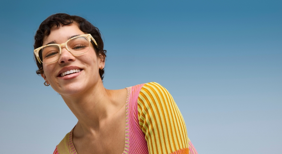 Woman wearing large, light-colored glasses and a striped, multicolored sweater, smiling against a blue background.