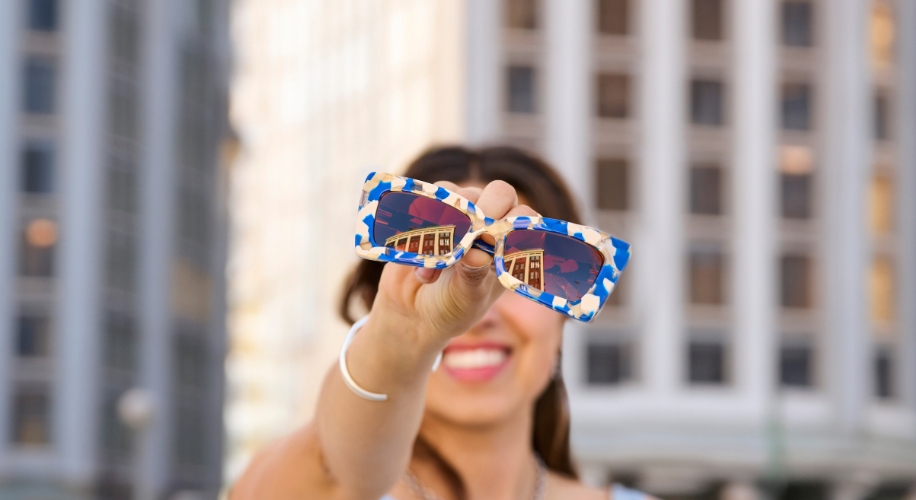 White and blue patterned sunglasses held up close. Background details of buildings are reflected in the lenses.