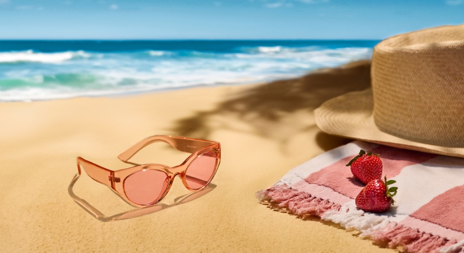 Pink sunglasses on sandy beach next to sun hat and two strawberries on a striped towel.