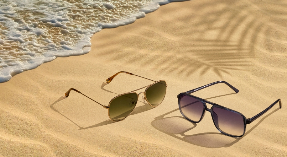 Two pairs of sunglasses on sand near the shoreline; one with green lenses, the other with gray lenses.