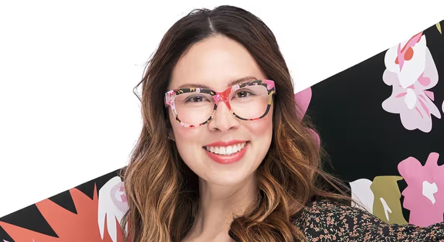 Woman wearing floral-patterned eyeglasses with pink and black colors, smiling in front of a floral background.