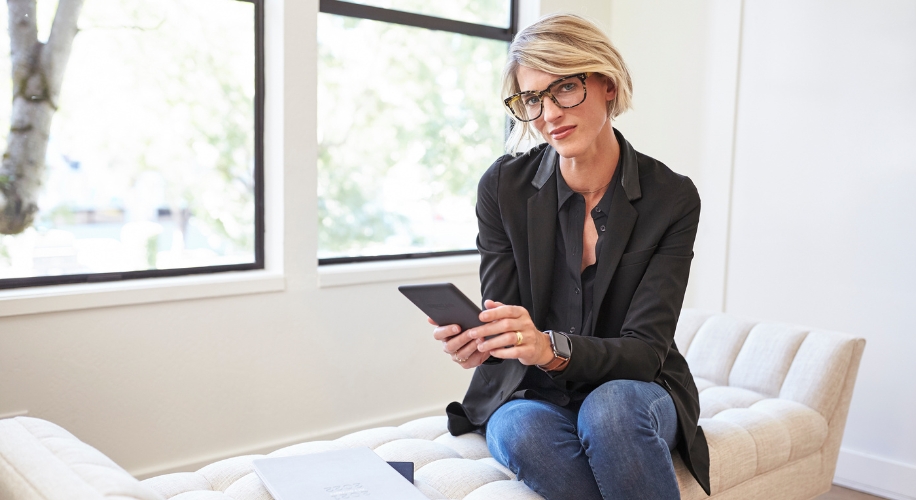 Woman holding a tablet while sitting on a cream-colored couch, wearing glasses and black business attire.