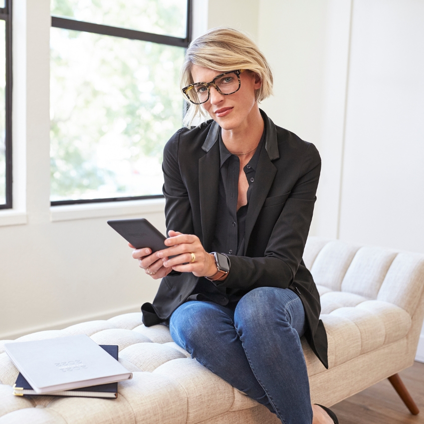 Person in a black blazer and jeans holding a black tablet. Two notebooks placed on a cushion seat.