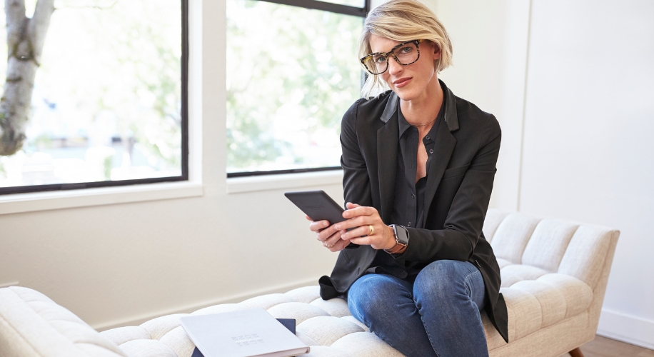 Woman wearing glasses holding an e-reader while sitting on a beige cushioned bench in a well-lit room.