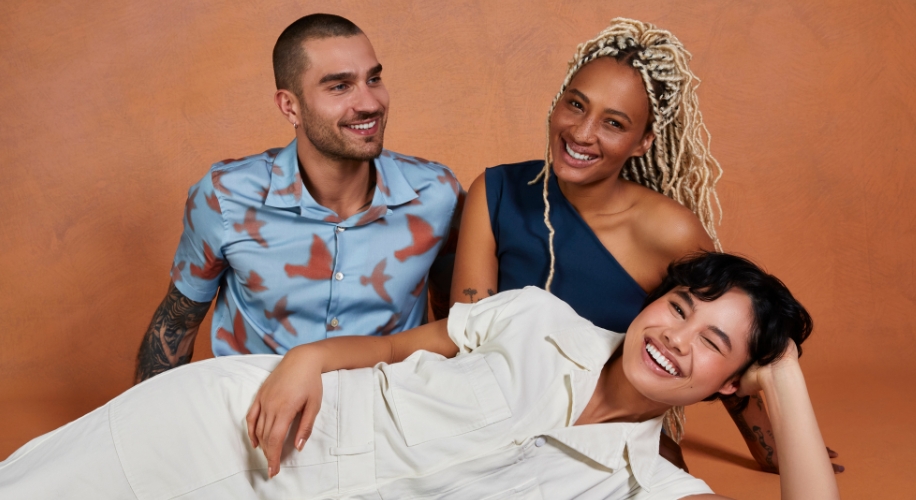 Three people wearing stylish, casual clothing, smiling and posing together against an orange background.