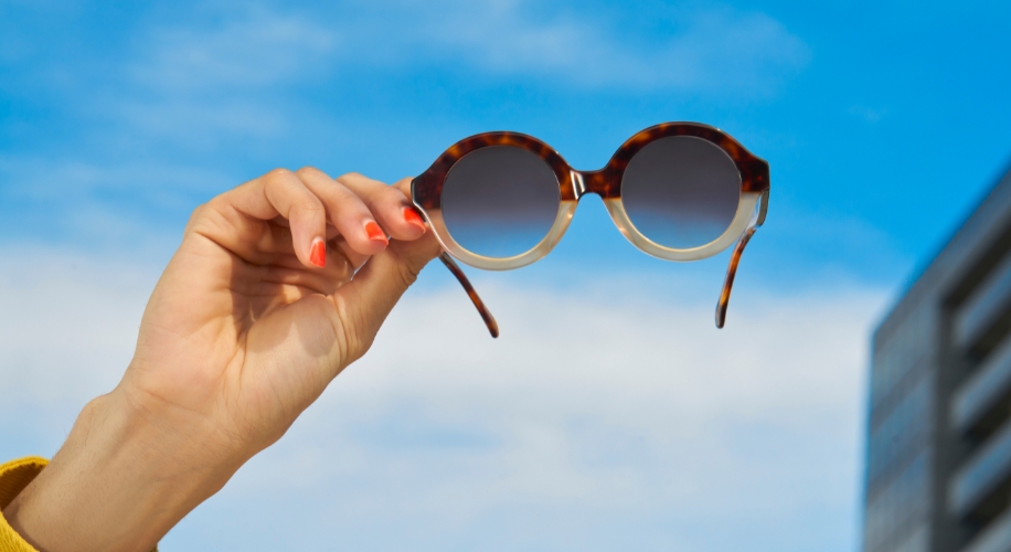 Round tortoiseshell sunglasses with gradient lenses held up by a hand against a blue sky background.