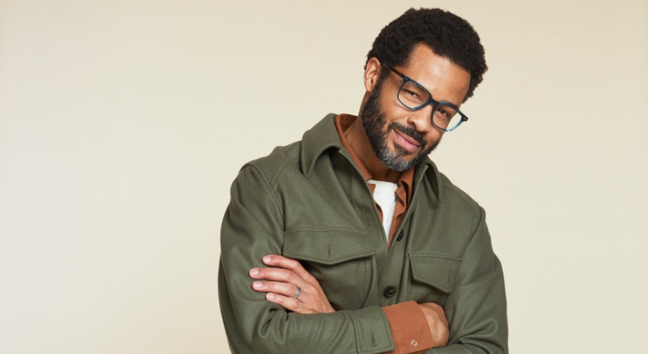 Man wearing an olive green jacket over a brown shirt, with black-framed glasses and arms crossed.