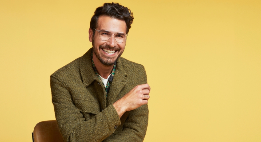 Man wearing clear glasses and a green tweed blazer, smiling in front of a yellow background.