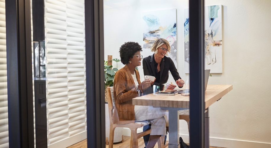 Two women collaborating at a wooden desk, one holding a coffee cup, the other pointing at a laptop.