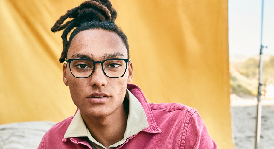 Person wearing black eyeglasses and a pink jacket with a yellow background.