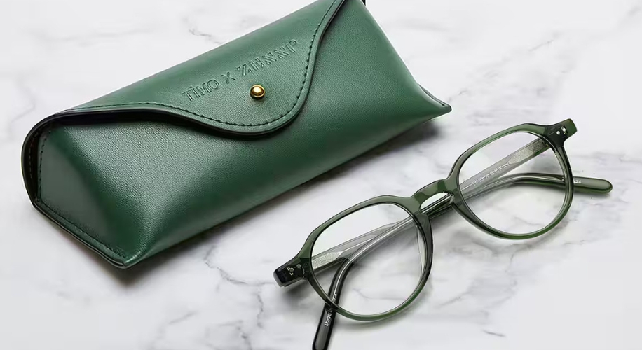 Green eyeglass case with gold button next to matching green round-frame eyeglasses. Text on case: &quot;TIM.O x ZAIBERT.&quot;