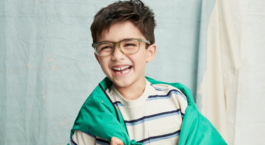 Smiling child wearing a striped shirt and green glasses, holding a green jacket over their shoulders.