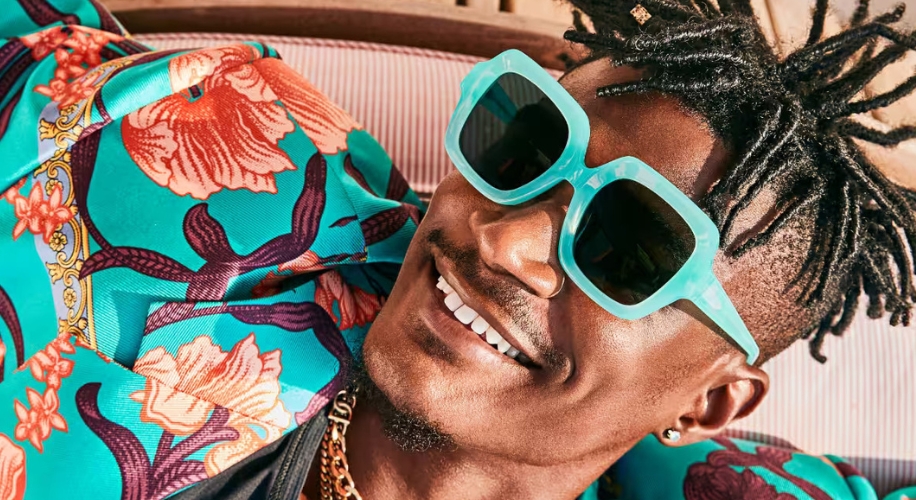 Man wearing oversized teal sunglasses and a vibrant floral-patterned shirt with a beaming smile.