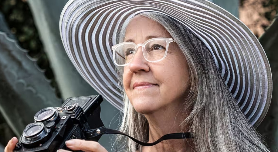 Woman holding a Yashica camera, wearing a striped wide-brimmed hat and glasses.