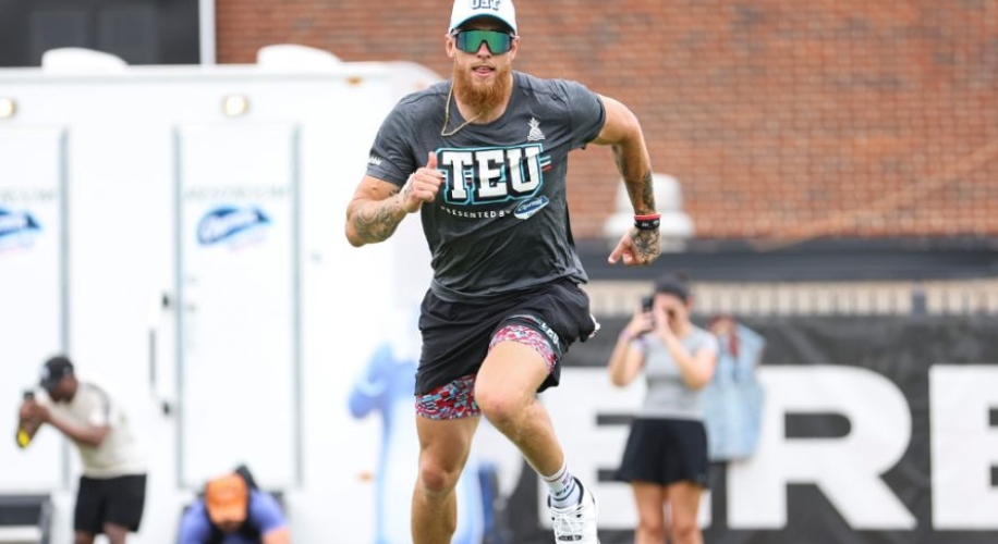 Man in motion wearing a grey &quot;TEU Presented by Old Spice&quot; t-shirt, patterned shorts, and a white cap with sunglasses.