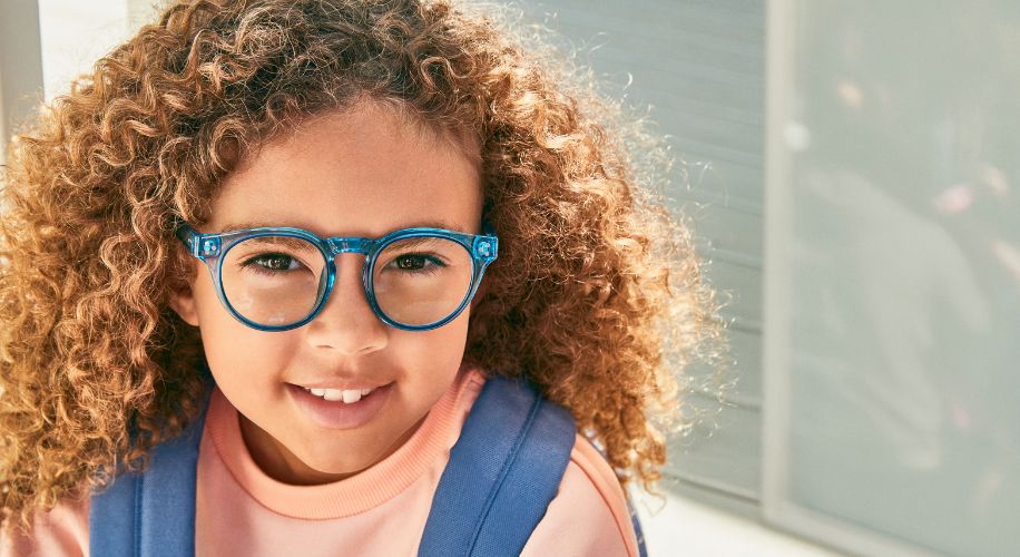 Child wearing blue eyeglasses with curly hair and a pink top.