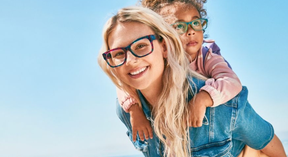 Two people wearing colorful eyeglasses, with one person giving the other a piggyback ride, smiling.