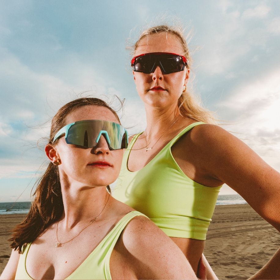 Two women with sunglasses; one wears blue-framed, reflective lenses, and the other, red wraparound glasses.