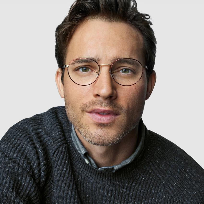 Thin, round, gold-rimmed eyeglasses worn by a person with short hair and light stubble, dressed in a grey sweater.