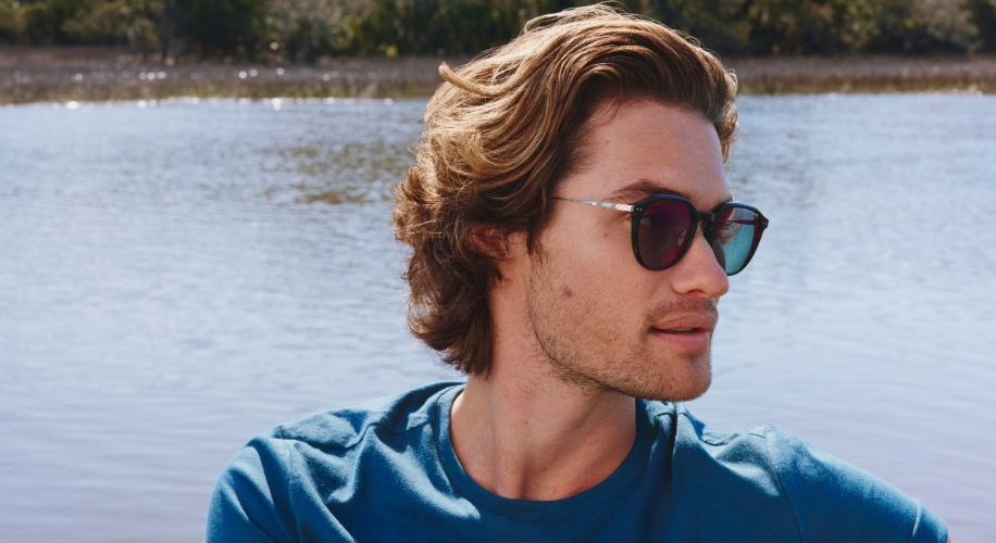 A person wearing sunglasses with reflective lenses and a blue shirt, standing by a body of water.