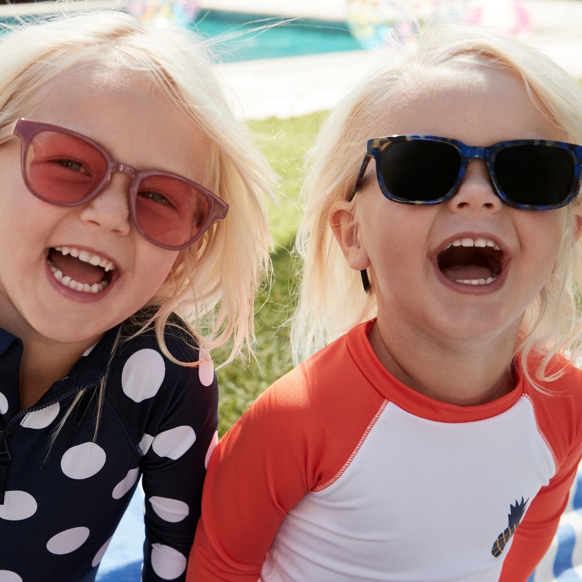 Kids wearing colorful sunglasses and long-sleeve rash guards with polka dot and pineapple designs, smiling.