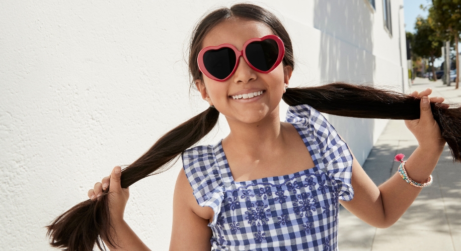 Girl wearing heart-shaped sunglasses and a blue checkered dress with embroidered flowers.