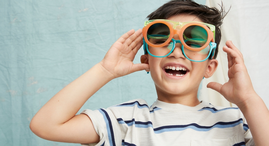 Child wearing two pairs of oversized, colorful sunglasses and laughing.