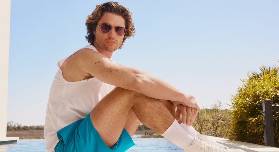 Man wearing white tank top, turquoise shorts, white socks, white sneakers, purple-tinted sunglasses.