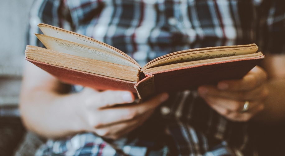 Person holding an open book with a red cover and yellowed pages.