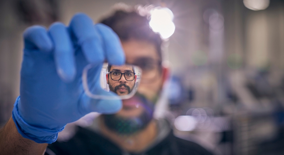 Person holding a transparent rectangular piece of glass with their face reflected in it, wearing blue gloves.