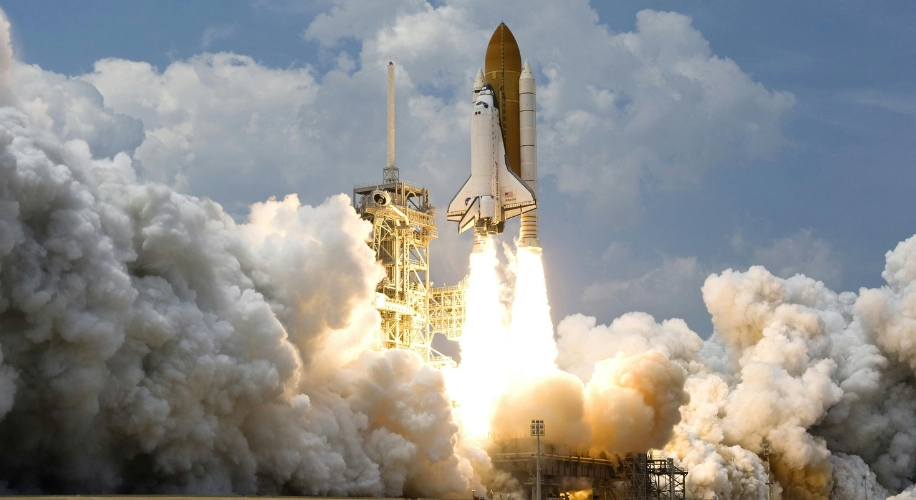 Space shuttle launching with powerful exhaust flames and smoke against a backdrop of clouds and blue sky.