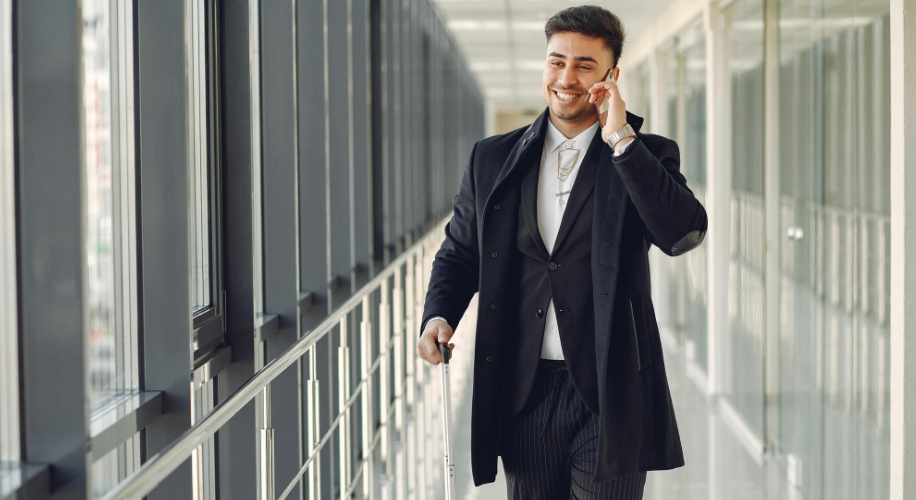 Man in a long black coat with a suitcase, smiling and talking on the phone.