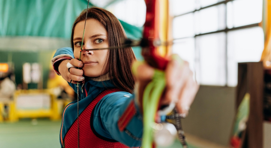 Archer aiming a bow, drawing the string back with intense focus. Wearing blue and red archery gear.