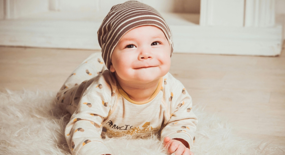 Baby wearing a striped hat and patterned pajamas, lying on a soft white rug, smiling cheerfully.