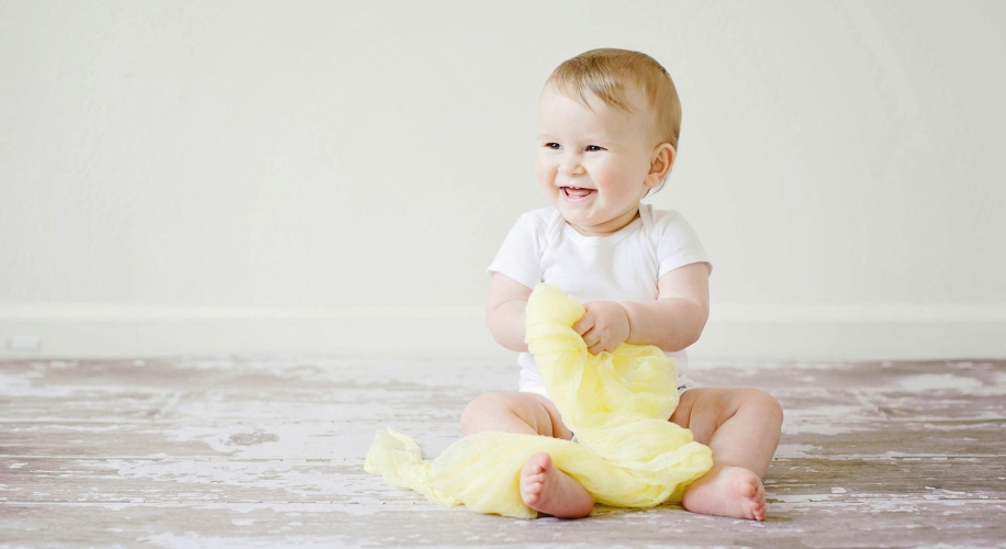 Smiling baby wearing a white onesie, holding a yellow cloth, seated on a light-colored wooden floor.