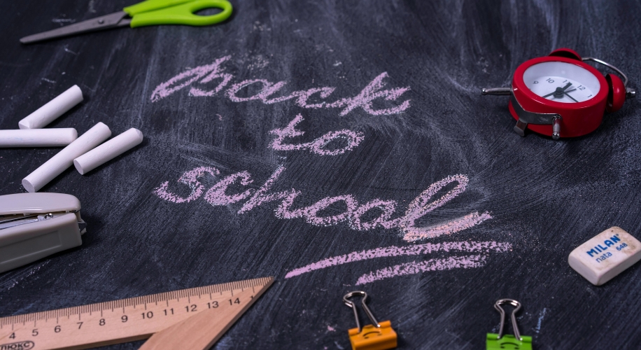 Back to school written in pink chalk on a blackboard with school supplies around it.