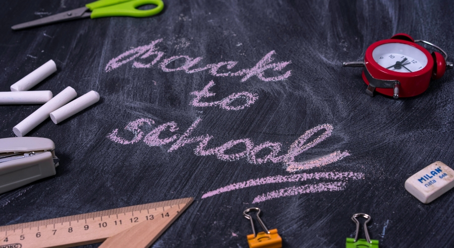 Back to school written in pink chalk on a blackboard with school supplies around it.