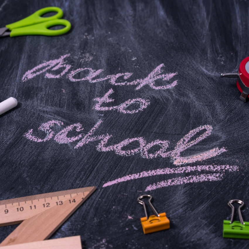 Back to school written in pink chalk on a blackboard, surrounded by scissors, a ruler, and binder clips.