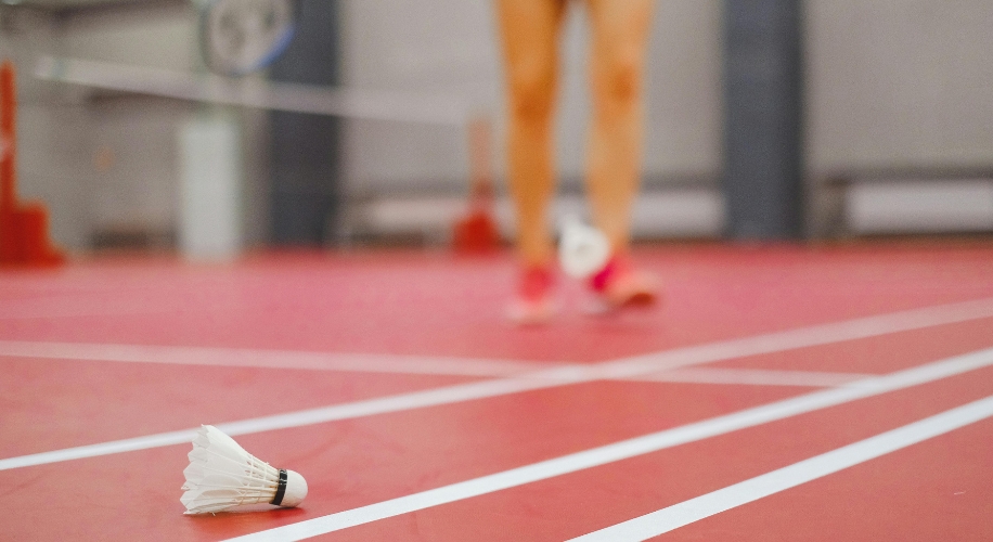 Shuttlecock on a red badminton court with white lines, blurred figure of a player in the background.