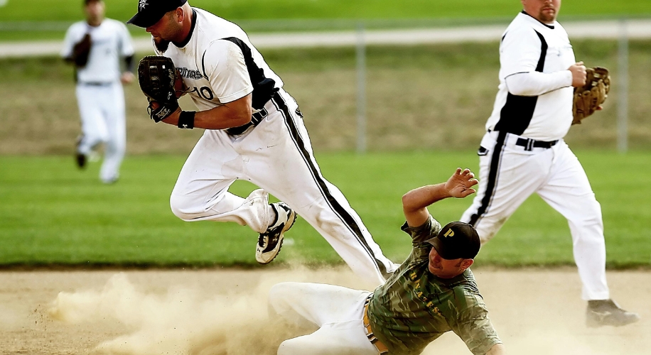 Baseball player in white and black uniform jumps to avoid player in camouflage while holding ball.