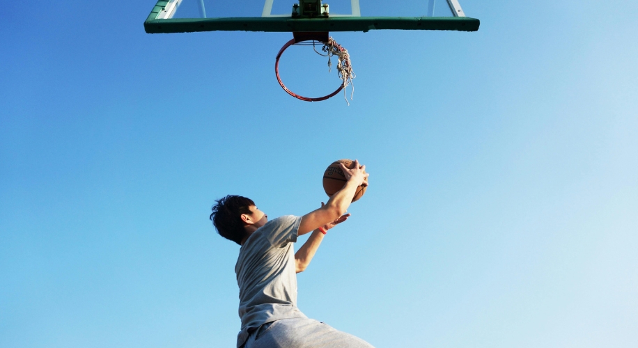 Person shooting a basketball towards a hoop with a broken net under a clear blue sky.