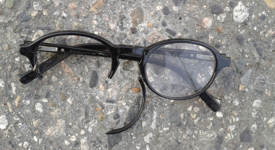 Broken black-framed glasses with one detached temple arm on a textured concrete surface.