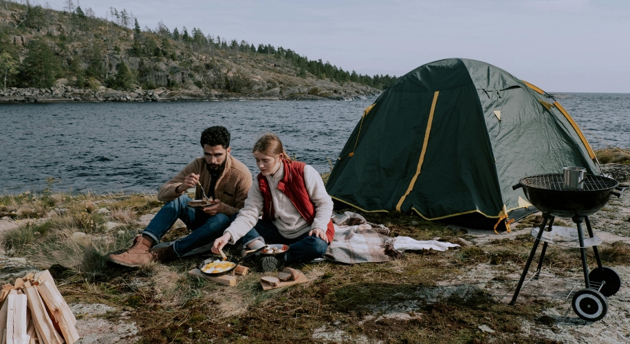 Campers cooking a meal next to a green tent by a lakeside.